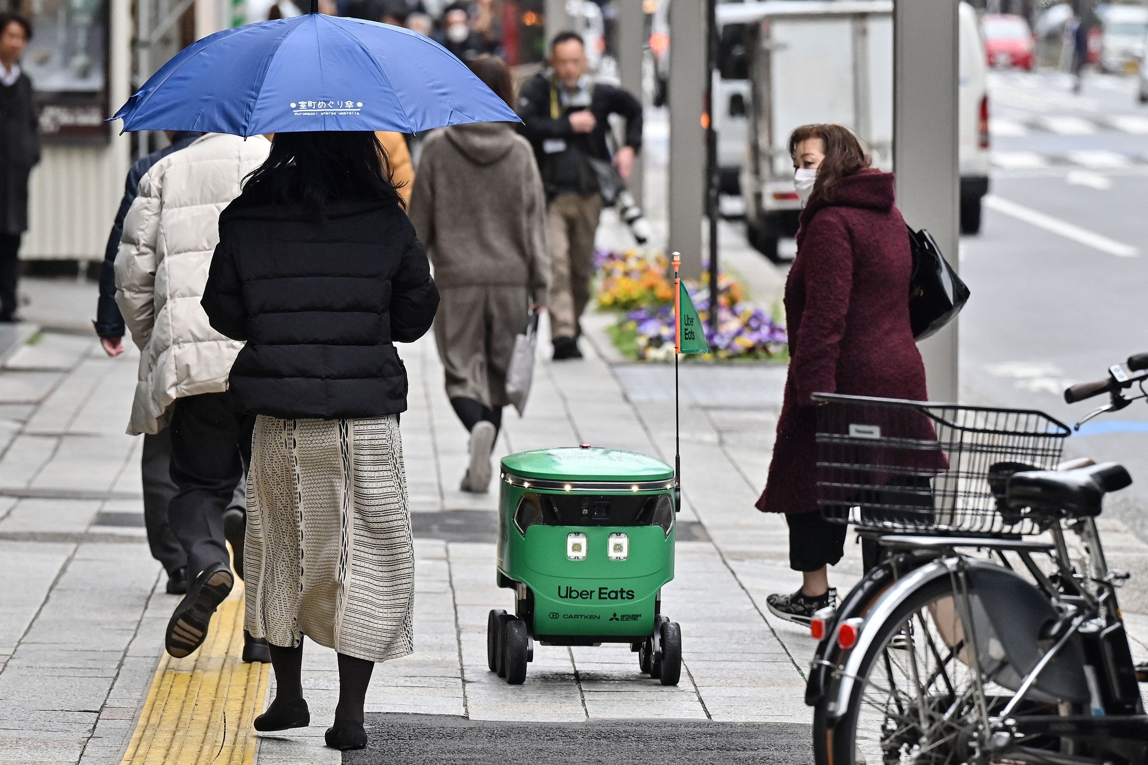 Peatones caminan junto a un robot no tripulado (c) mientras navega por una calle durante una demostración de un servicio de entrega con robots realizado por Uber Eats Japan, Mitsubishi Electric y el desarrollador de robots Cartken en el centro de Tokio el 5 de marzo de 2024. 