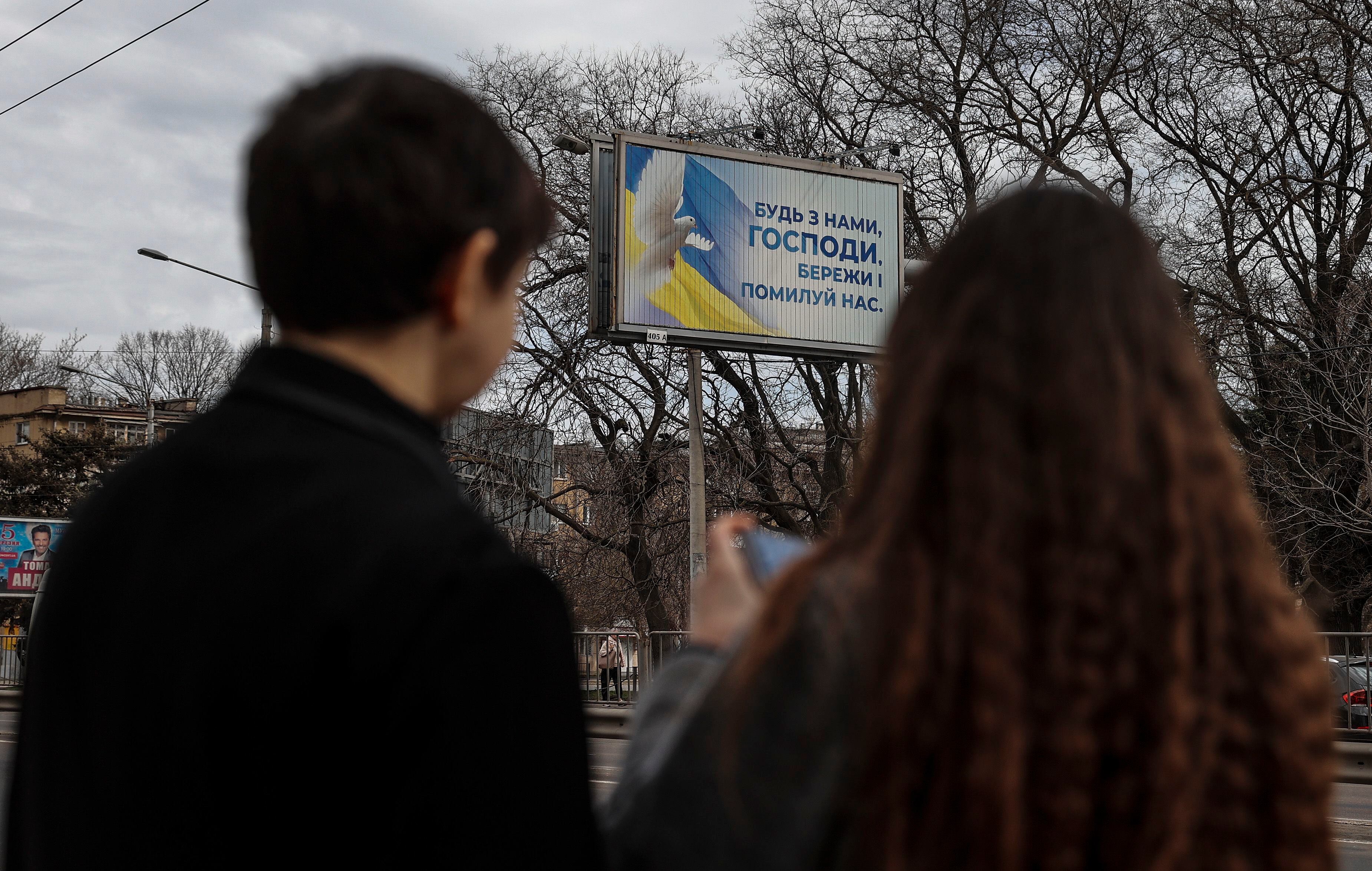 Dos personas permanecen paradas ante un cartel en el que se puede leer "Dios está con nosotros, sálvanos y ten piedad" y que está situado en una calle de Odesa (Ucrania). EFE/Manuel Bruque 